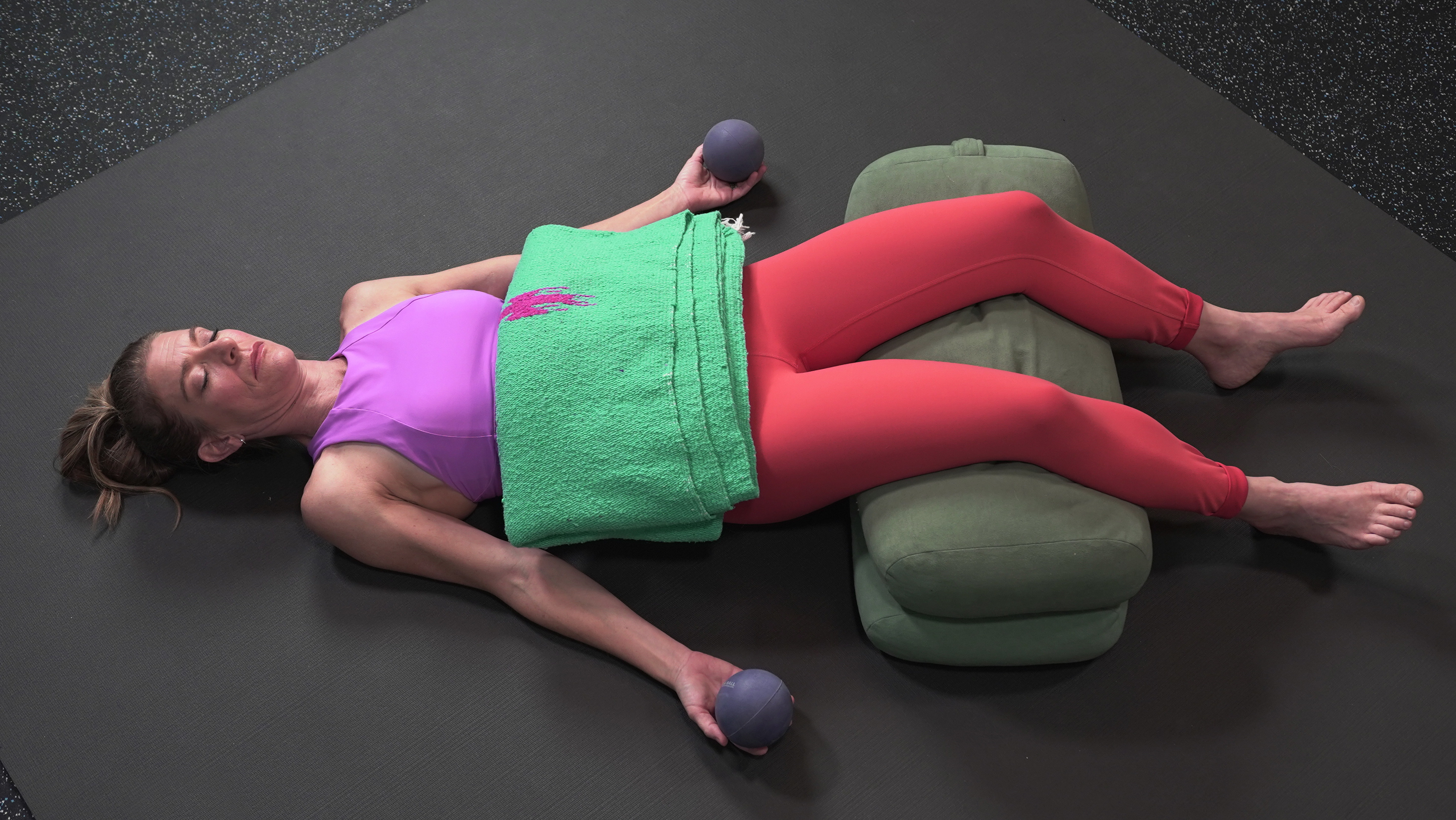 Woman lies in constructive rest position; two yoga bolsters propping up knees, a folded blanket on the abdomen and alpha self massage balls in hand to add weight and comfort